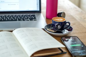 Tisch, auf dem sich diese Gegenstände befinden: aufgeklappter Laptop, Kalender, Tasse Kaffee mit Untertasse sowie ein Smartphone.