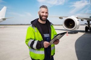 Mann mit Klemmbrett in der Hand steht auf Start- und Landebahn eines Flughafens. Im Hintergrund befinden sich zwei Flugzeuge, die teilweise zu sehen sind.