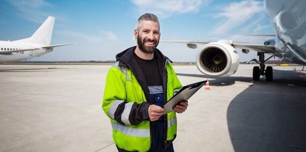 Mann mit Klemmbrett in der Hand steht auf Start- und Landebahn eines Flughafens. Im Hintergrund befinden sich zwei Flugzeuge, die teilweise zu sehen sind.