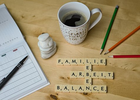 Holzplatte, auf der sich diese Dinge befinden: Tischkalender, schwarzer Kugelschreiber, Tasse mit Kaffee, drei Bleistifte in den Farben grün, orange und rot sowie ein Schriftzug "Familie-Leben-Arbeit-Balance" in Form von Dominosteinen.