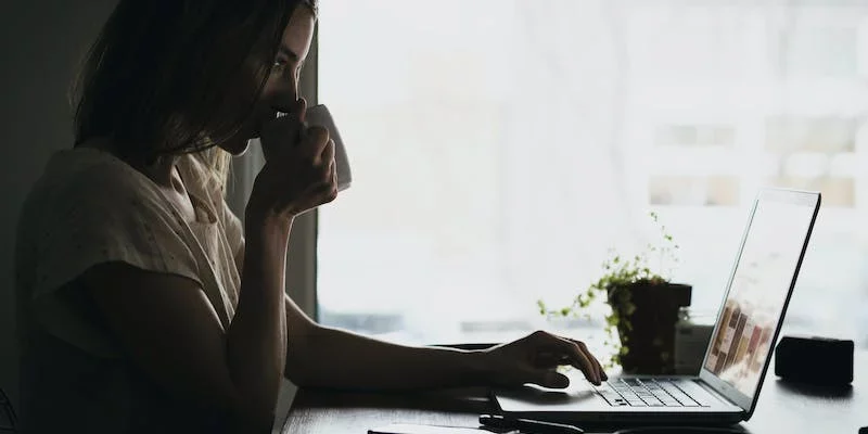 Frau sitzt in einem dunklen Raum mit einem Kaffe an ihrem Laptop