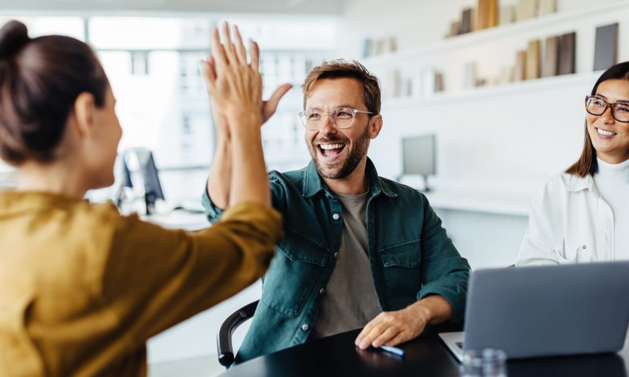 Drei Mitarbeiter, zwei Frauen und ein Mann, sitzen an einem Konferenztisch, auf dem ein Laptop steht. Der Mann und eine Frau machen einen High Five-Handschlag.