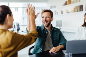 Drei Mitarbeiter, zwei Frauen und ein Mann, sitzen an einem Konferenztisch, auf dem ein Laptop steht. Der Mann und eine Frau machen einen High Five-Handschlag.