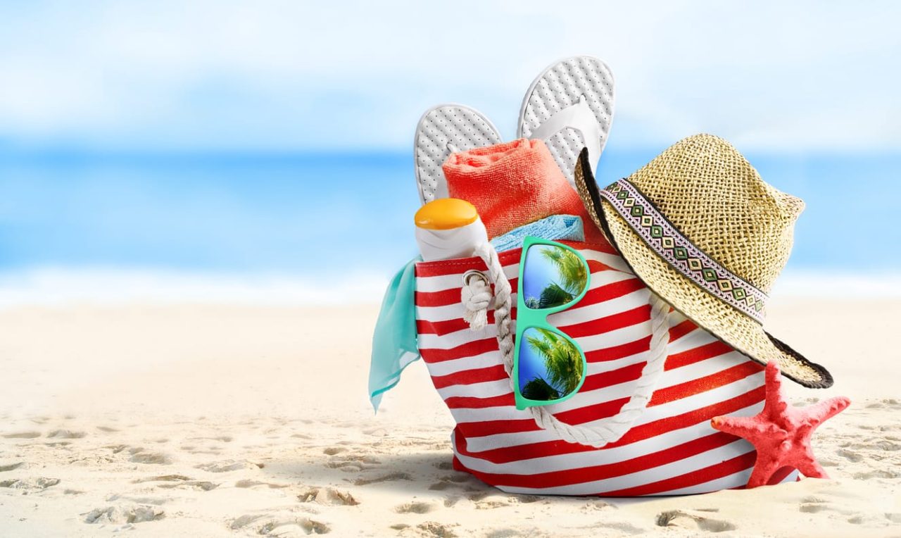 Rot-weiß gestreifte Tasche mit Hut, Sonnenbrille, Sonnenmild, Handtuch und Badeschuhen steht an einem Strand