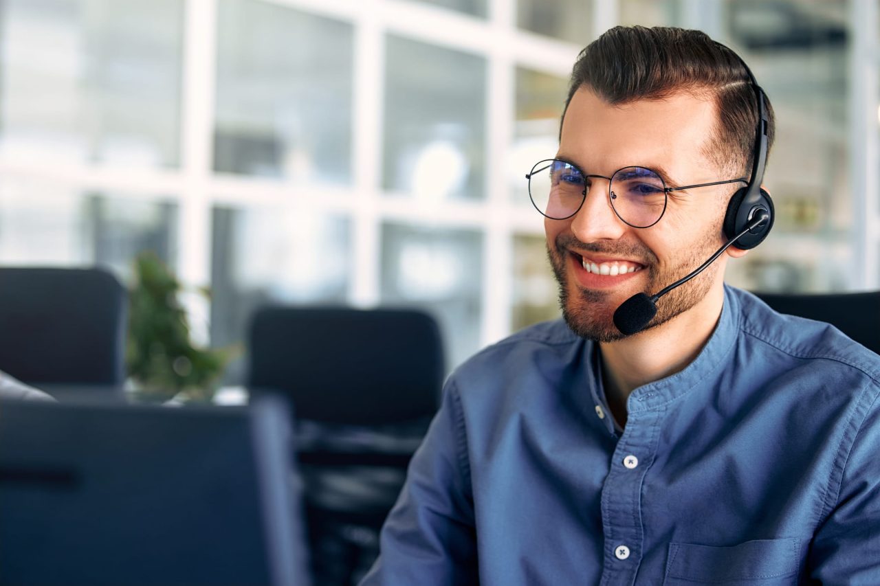 Lächelnder junger Mann mit Brille und Headset sitzt vor einem PC. Der Hintergrund ist verwischt