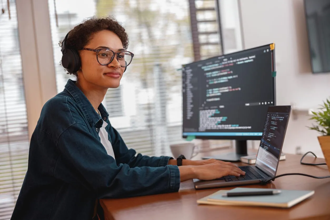 Junge Programmiererin mit Kopfhörern und Brille arbeitet lächelnd an Laptop und großem Bildschirm mit Programmiercode in modernem Büro.