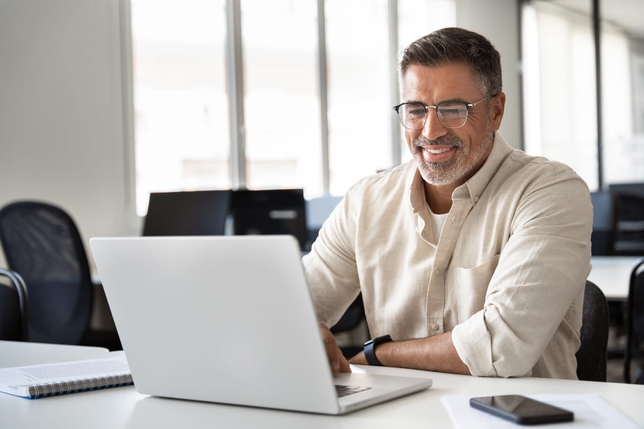 Mittelalter Mann mit Brille und grauem Bart sitzt lächelnd in einem modernen Büro vor einem Laptop. Er trägt ein beiges Hemd, im Vordergrund liegt ein Smartphone und ein Notizbuch.