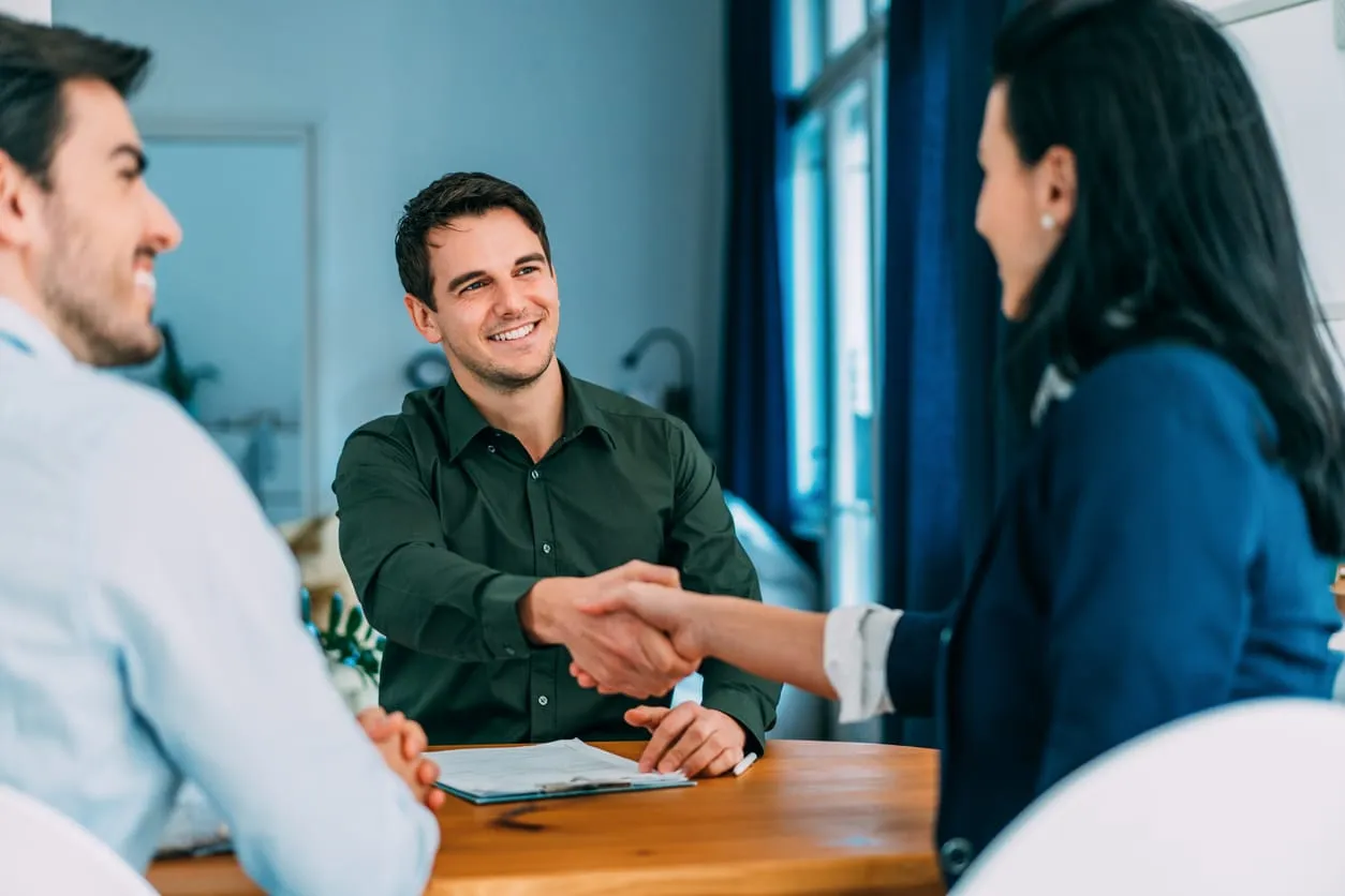 Drei Personen in einem Vorstellungsgespräch: Ein Bewerber in dunklem Hemd lächelt und schüttelt der gegenüber sitzenden Frau die Hand, ein weiterer Mann sitzt daneben. Freundliche Atmosphäre in einem modernen Büro.