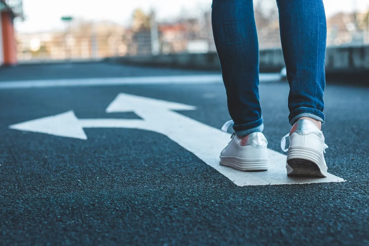 Eine Person in Jeans und weißen Sneakern steht auf einem Asphaltweg vor einem Richtungspfeil, der sich in zwei gegensätzliche Richtungen verzweigt. Das Bild symbolisiert eine Entscheidungssituation oder eine berufliche Neuorientierung