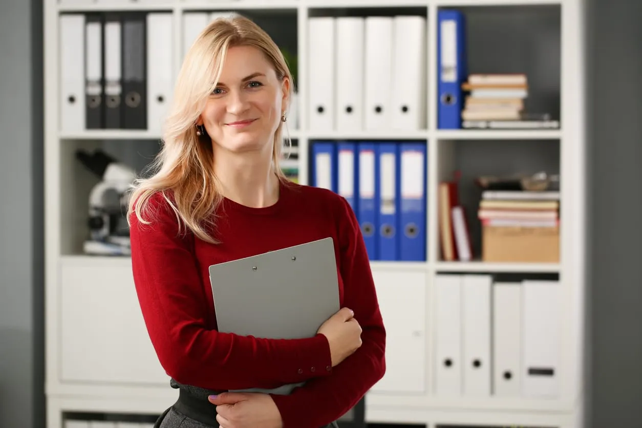 Freundliche Geschäftsfrau mit langen blonden Haaren und rotem Pullover steht mit einem Klemmbrett im modernen Büro vor einem Regal mit Aktenordnern und Büchern.