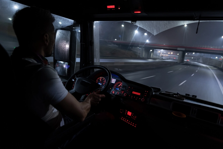 Ein Lkw-Fahrer steuert sein Fahrzeug konzentriert auf einer nächtlichen, beleuchteten Autobahn. Die Perspektive zeigt das beleuchtete Armaturenbrett, die Straße und die Brücke im Hintergrund, wodurch die Stimmung einer typischen Nachtschicht vermittelt wird.