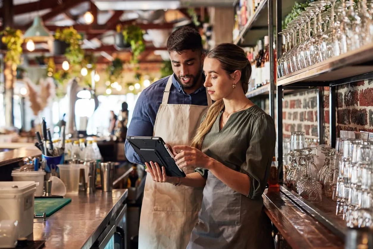 Zwei Mitarbeitende in einem Restaurant stehen hinter der Theke und schauen gemeinsam auf ein Tablet. Beide tragen Schürzen, im Hintergrund sind Gläser, Flaschen und Pflanzen zu sehen. Die Atmosphäre wirkt modern und einladend.