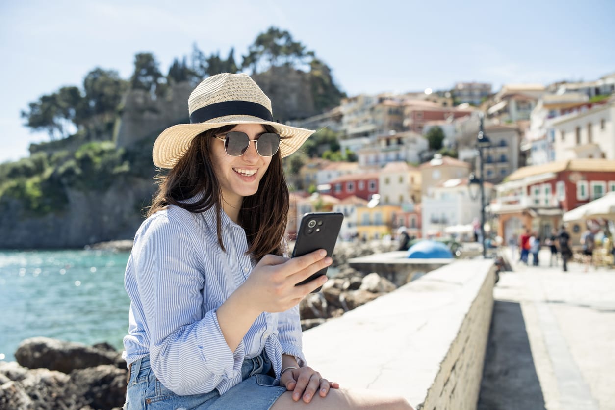Junge Frau mit Sonnenhut und Sonnenbrille sitzt lächelnd am Wasser und schaut auf ihr Smartphone. Im Hintergrund sind leicht verschwommen bunte Häuser an einem Hang in einem mediterranen Küstenort zu sehen.