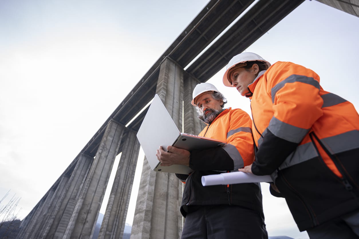 Zwei Bauingenieure in oranger Schutzkleidung prüfen auf einer Baustelle unter einer Brücke technische Daten auf einem Laptop.