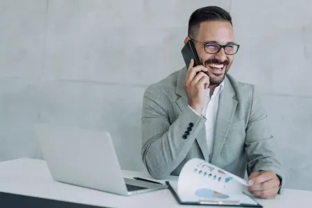 Lächelnder Geschäftsmann mit Brille und 3-Tage-Bart hält einen Telefonhörer in der Hand. Er lehnt seine Arme auf einen weißen Tisch, auf dem sich ein silberner Laptop und ein Klemmbrett liegen.