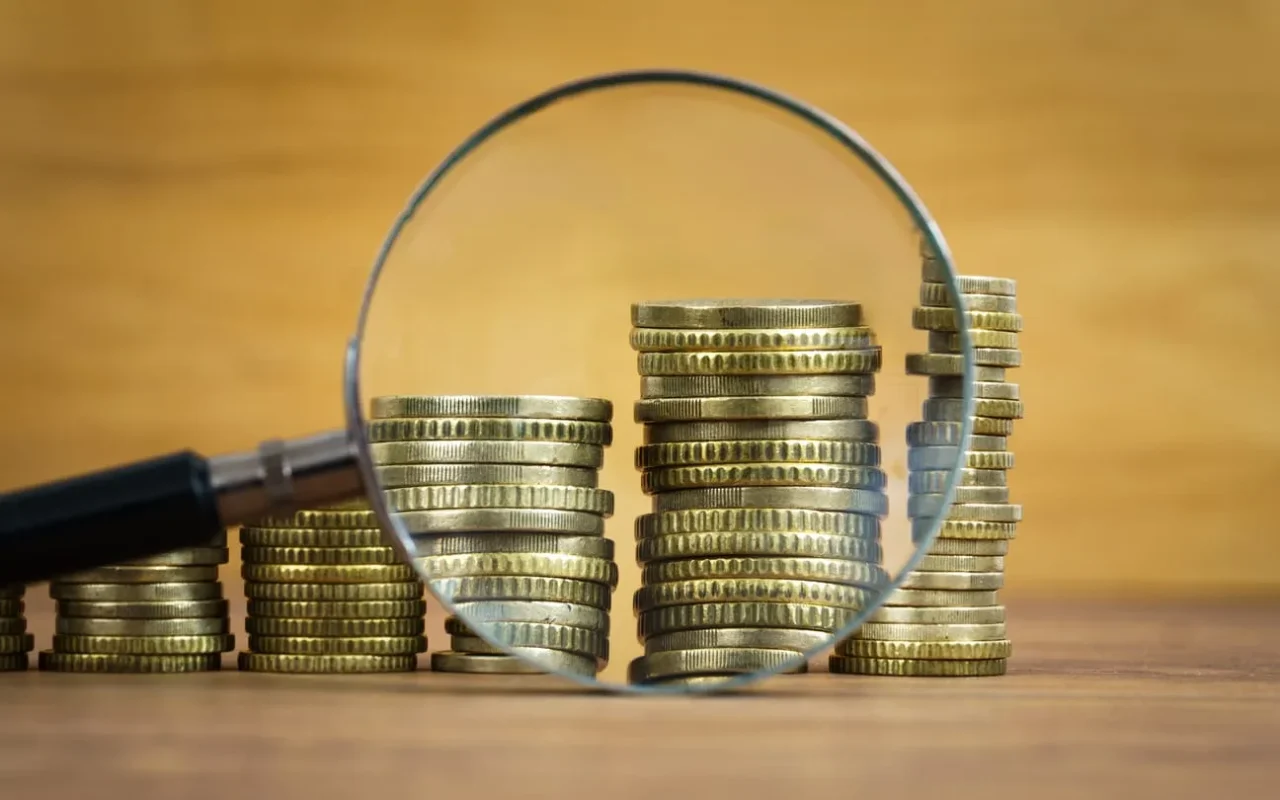 Symbolbild zur Gehaltstransparenz zeigt Münzstapel in Stellenanzeigen unter Lupe