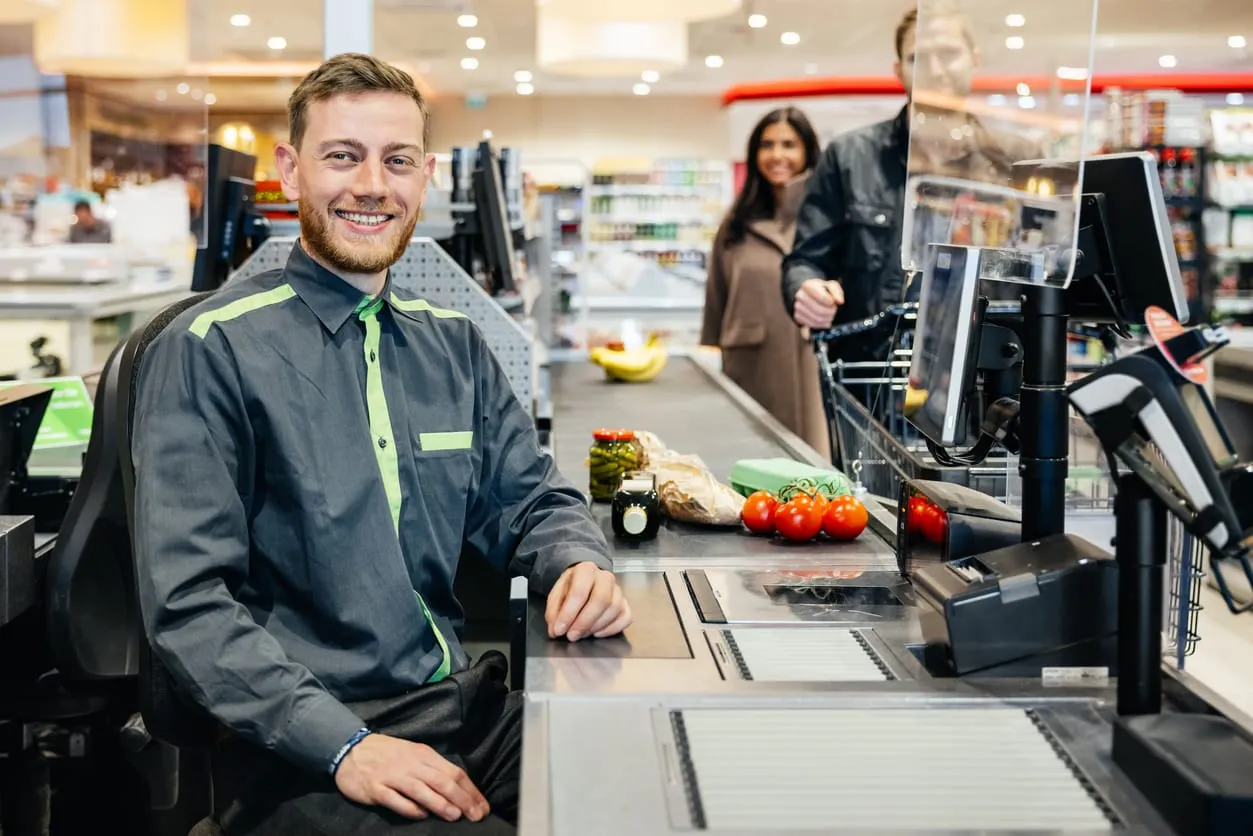 Junger lächelnder Verkäufer mit rotblonden Haaren und rotblondem Bart sitzt an einer Supermarktkasse. Auf dem Band liegen Waren. In der Schlange steht ein Mann mit Einkaufswagen und dahinter eine Frau.