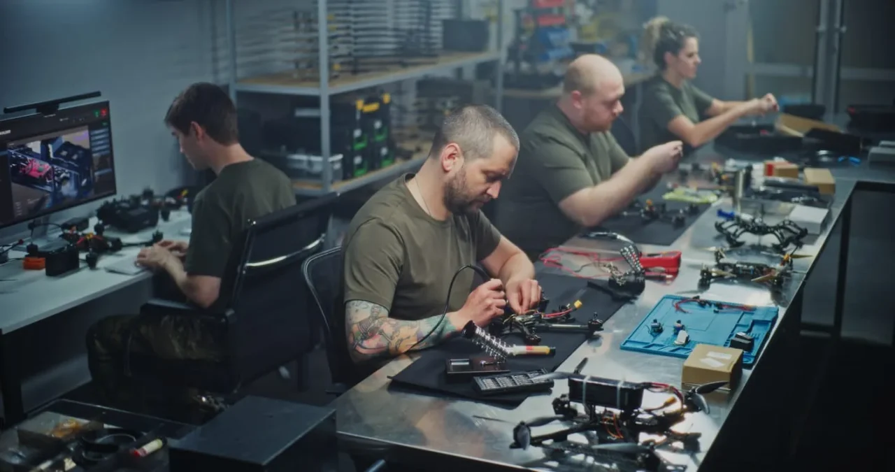 Personen in grünen Hemden sitzen an einem Tisch und werkeln an militärischen Geräten