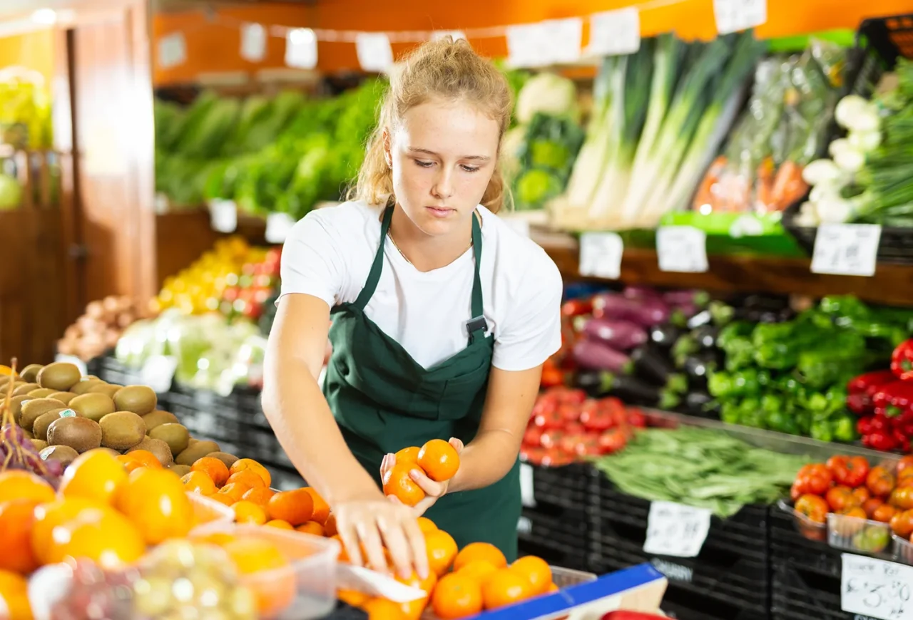 Junge blonde Verkäuferin mit weißem T-Shirt und dunkelgrüner Schürze steht in der Gemüseabteilung eines Supermarktes, hat drei Mandarinen in der Hand und blickt auf Mandarinen-Auslage